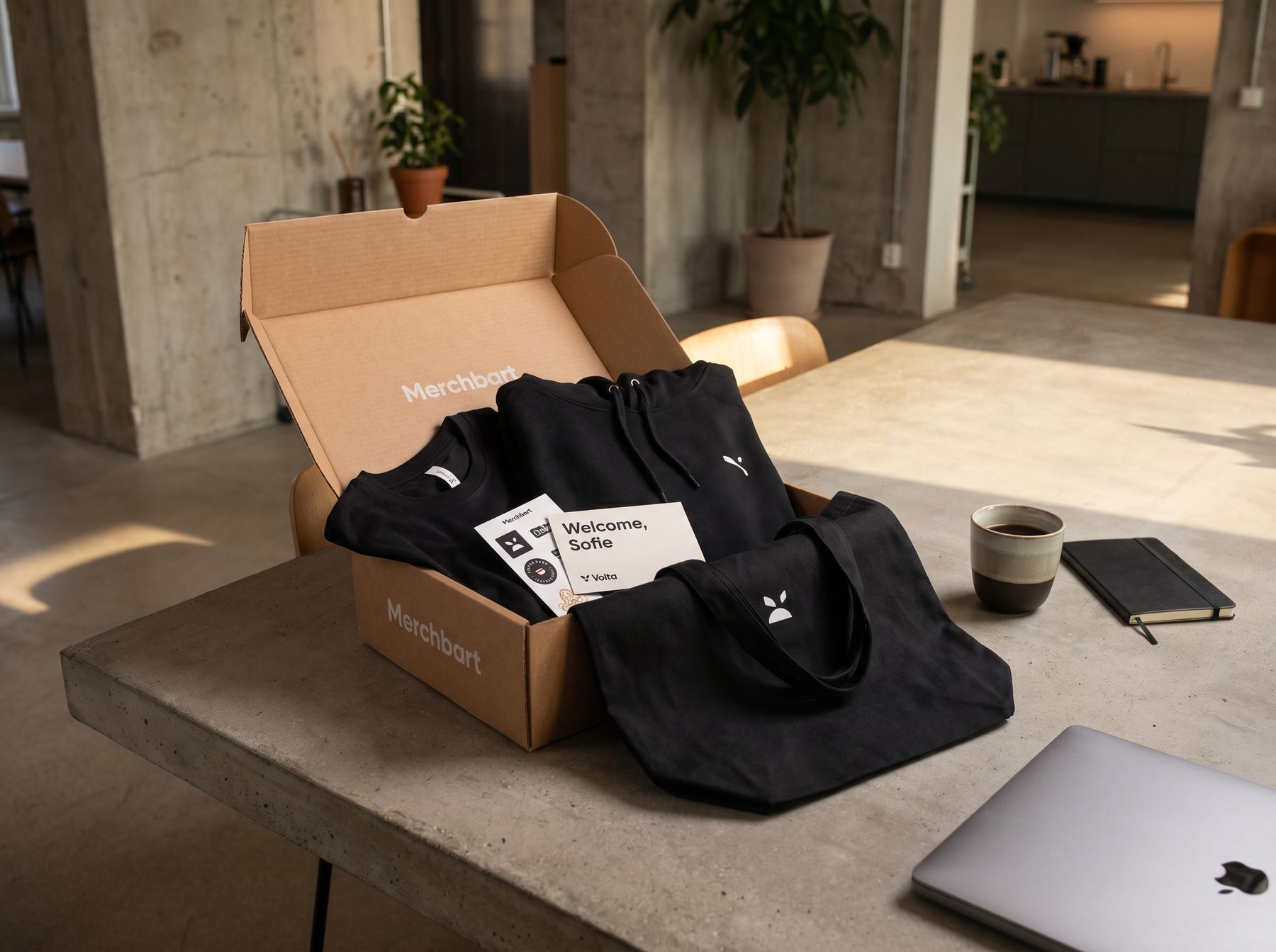 Merchbart velkomstpakke på skrivebord med hoodie, tote, stickers og et 'Velkommen, Sofie'-kort fra Volta